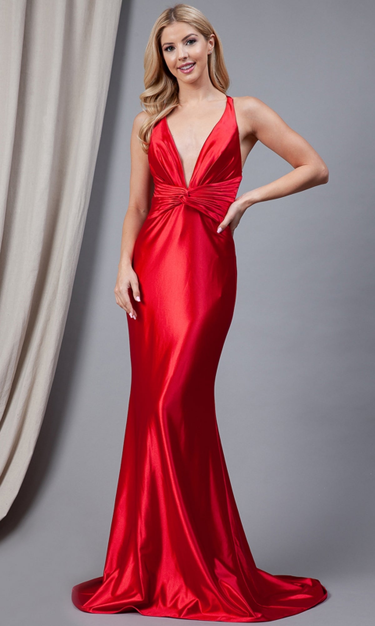 A woman models the Twist-Waist Deep V-Neck Long Prom Dress 5039, a sleeveless floor-length red gown, in front of a gray backdrop and a beige curtain.